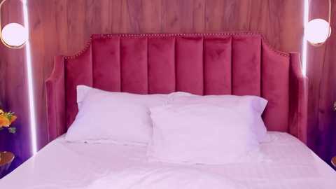 Media: Video of a plush, white bed with a tufted, deep pink headboard against wooden paneled walls, featuring two bedside lamps with spherical white shades.