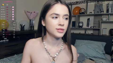 Media: Video of a young woman with fair skin and long brown hair, wearing a beaded necklace, in a bedroom with a gray bed, a TV, and a shelf with books.