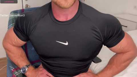 Media: Video of a muscular man in a black Nike shirt, with visible tattoos on his forearms and a watch on his left wrist, sitting on a blue and green plaid chair in a white room.