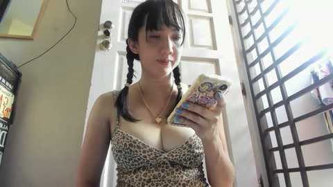 Media: Video of a fair-skinned woman with twin braids, wearing a leopard-print top, taking a selfie in a sunlit room with a door and screen in the background.