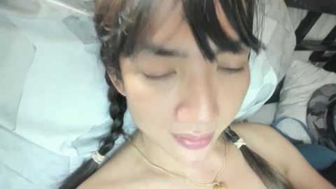 Media: Video of an Asian woman with straight black hair in braids, wearing a gold necklace, lying on a white pillow in a dimly lit room.