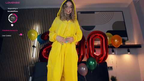 Media: Video of a smiling woman in a yellow raincoat and hood, surrounded by colorful balloons in a modern living room with a large \"Happy Birthday\" sign.