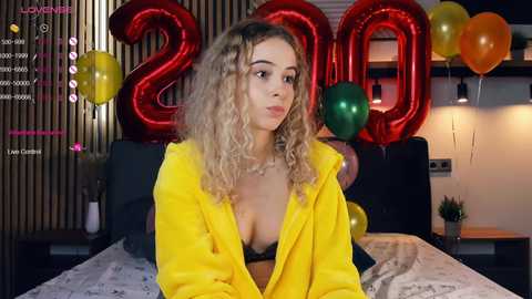 Media: A video of a young woman with curly blonde hair, wearing a yellow jacket and black bra, sitting on a bed adorned with \"2020\" balloons.