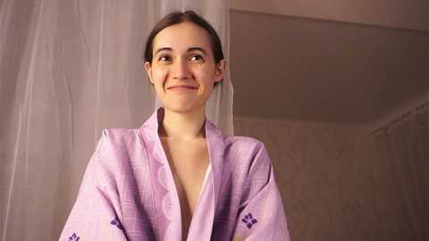 Media: Video of a smiling young woman with light skin and brown hair in a pale lavender robe, standing in a softly lit room with white curtains and textured beige walls.