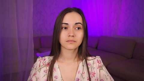 Media: Video of a young woman with long, straight brown hair and light skin, wearing a floral blouse, indoors with purple lighting, soft focus background of a beige couch.