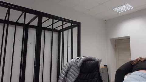 Media: Video of a bare, rectangular room with a metal-framed cage on the left and a woman in a gray puffer jacket and black shirt standing in the center, with a light blue wall and fluorescent ceiling lights.