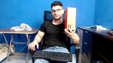 Media: Video of a young man with short brown hair, wearing glasses and a black t-shirt, sitting at a desk, holding a phone in front of a blue wall, with a messy workspace.