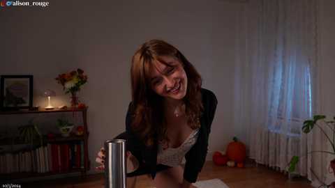 Media: Video of a smiling, brown-haired woman in a black cardigan and white lace bra, pouring tea from a kettle, in a cozy, dimly-lit living room with autumn decorations.