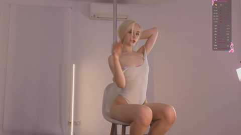 Media: A video of a slender, blonde woman in a sheer white bodysuit, sitting on a chair, arms raised, in a minimalist, dimly-lit room with an air conditioner and a pink poster on the wall.