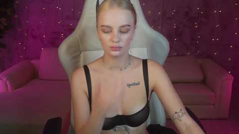 Media: Video of a fair-skinned, blonde woman with tattoos, wearing a black bra, sitting on a white gaming chair in a dimly lit room with pink and purple lighting.