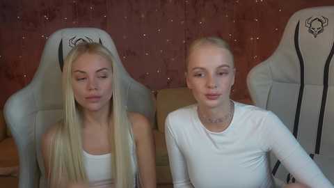 Media: Video of two young women with long blonde hair, wearing white tank tops, sitting in gaming chairs against a textured, reddish-brown wall.