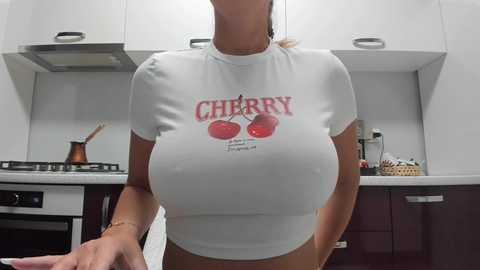 Media: Video of a woman in a modern kitchen, wearing a tight, white crop top with \"CHERRIES\" and a cherry graphic, showcasing her large breasts. Background features white cabinets, a stove, and utensils.