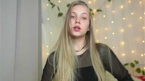 Media: Video of a young Caucasian woman with long blonde hair, wearing a sheer black top, standing in a softly lit room with fairy lights and green foliage in the background.