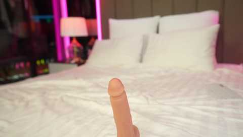 Media: Video of a hand holding an erect, circumcised penis on a neatly made bed with white sheets, two pillows, and a headboard. The background features a modern bedroom with purple and pink neon lights and a nightstand with a lamp.