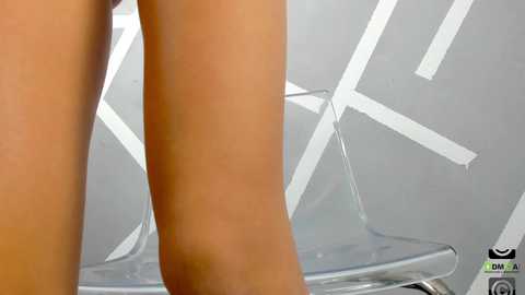 Media: Video of a close-up of a woman's bare, smooth, tanned legs on a transparent, modern, white chair with a geometric design. The background features a gray wall with white geometric patterns.