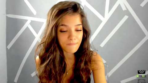 Media: Video of a young woman with long, wavy brown hair, closed eyes, and a serene expression, against a modern, geometric grey and white background.