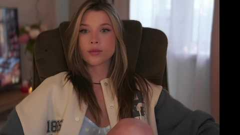 Media: Video of a young woman with long blonde hair, wearing a white lab coat over a grey sweater, sitting in a brown chair, looking thoughtful.