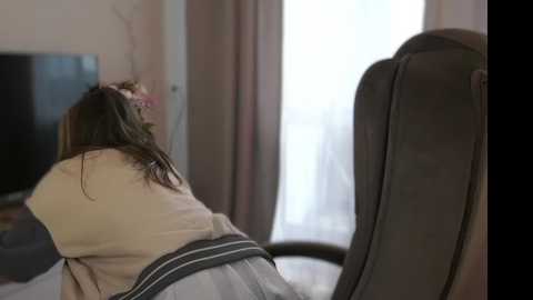 Media: Video of a woman with long brown hair, wearing a beige sweater and gray skirt, seated in a gray office chair, facing a large TV screen on a beige wall, in a dimly lit room.