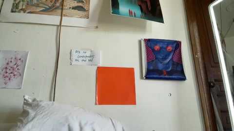 Media: Video of a bedroom wall with a white background, featuring a colorful, abstract painting, an orange square, and a handwritten note; a framed artwork and a window frame are visible in the background.