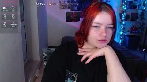 Media: A video of a fair-skinned woman with shoulder-length red hair, wearing a black shirt, sitting pensively indoors with a blue-tinted background, displaying a digital temperature readout.