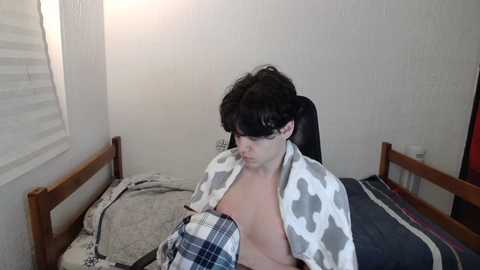 Media: A video of a young, fair-skinned man with messy black hair, wearing a cow-patterned blanket, lying on a bed with plaid and gray sheets, in a dimly lit, plain room.