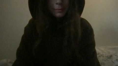 Media: Video of a person with long, dark hair wearing a hooded, dark coat, partially obscuring their face, in a dimly lit, beige-toned room. The image is grainy and slightly blurred, giving it a moody, mysterious atmosphere.