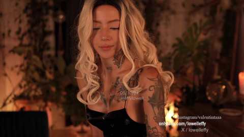 Media: Video of a smiling, tattooed, blonde woman with wavy hair, wearing a black top, in a dimly lit room with plants, candles, and a fireplace in the background.