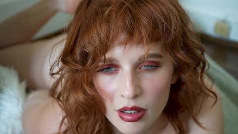 Media: Video of a fair-skinned woman with wavy red hair, blue eyes, and bold red lipstick, lying on a white surface, showcasing a close-up of her face.