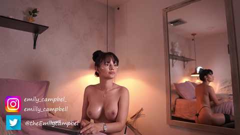 Media: A video shows a topless Asian woman with dark hair in a bun, sitting on a bed, looking at her reflection in a mirror. The room is dimly lit with a warm glow.