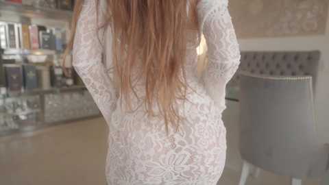 Media: Video of a woman with long, straight, light brown hair, wearing a white, lace, long-sleeved dress, standing with hands behind her back in a modern, brightly lit store with shelves of products in the background.