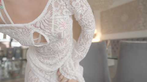 Media: A close-up video of a woman wearing a delicate, white lace dress with floral patterns. Her hands are on her hips, revealing her slender waist. The background shows a blurred, cozy living room with soft lighting and patterned wallpaper.