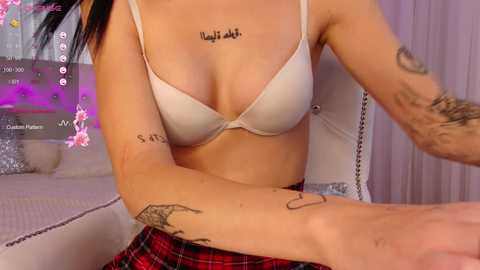 Media: Video of a light-skinned woman with black hair, wearing a beige bra, plaid skirt, and visible tattoos, seated on a white chair in a bedroom with purple curtains and a TV screen.