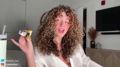 Media: Video of a young woman with curly, light brown hair, wearing a white robe, holding a condom wrapper, indoors with a flat-screen TV and potted plant in the background.