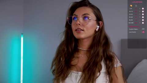 Media: Video of a young woman with long, wavy brown hair, wearing clear glasses and a white top, standing in a dimly lit room with a glowing blue light on the left.