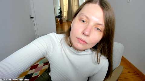 Media: A video of a young Caucasian woman with fair skin and straight brown hair, wearing a white ribbed sweater, sitting on a wooden floor with a colorful rug in the background.