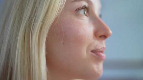 Media: Video of a close-up side profile of a young Caucasian woman with light skin and straight, platinum blonde hair. Her expression is serene, with a single tear on her cheek, hinting at emotional depth. The background is softly blurred, emphasizing her face.