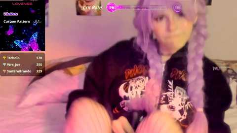 Media: Video of a young woman with lavender hair, wearing a black hoodie with white skull graphics, lying on a bed with a pink pillow. Background shows a virtual chat screen.