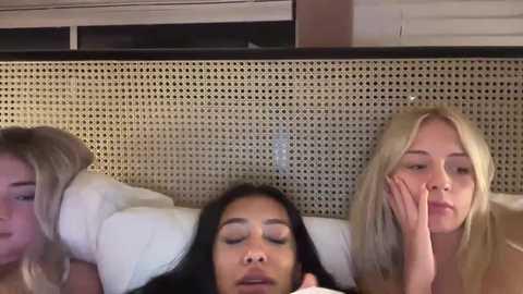 Media: Video of three young women with varying skin tones and hair colors, lying on white pillows, looking tired. Background features a beige perforated panel.