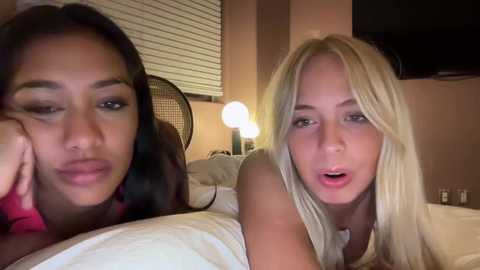 Media: Video of two women lying on a bed, one with dark hair and the other with blonde hair, both looking at the camera with slightly open mouths.