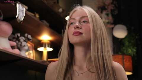 Media: Video of a young, fair-skinned blonde woman with long straight hair, wearing a strapless top, in a warmly lit bar. Background includes shelves, a lit lamp, and dimly visible decorations.