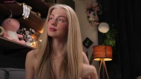 Media: A video of a young, blonde woman with fair skin and a slim physique, topless, standing in a dimly lit room filled with plush toys, a lamp, and a plant.