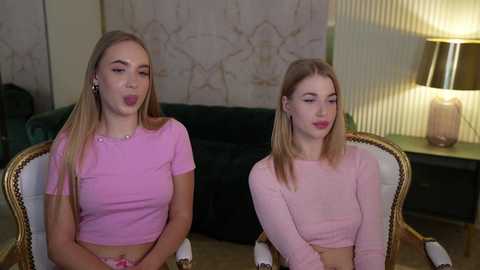 Media: Video of two young, blonde, fair-skinned women in matching pink crop tops, sitting on a dark green velvet sofa, making silly faces. Background features a beige, floral-patterned wall and a modern, beige lamp.