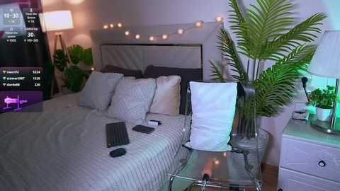 Media: Video of a modern bedroom with a queen-sized bed, beige bedding, and a white chair with a white cushion. A potted plant, a lamp, and a TV screen with a fitness app are visible.