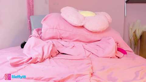 Media: Video of a pink bed with a plush white Hello Kitty pillow and a messy, wrinkled pink duvet. The room has a soft, pastel color palette, featuring a small vase of dried flowers.