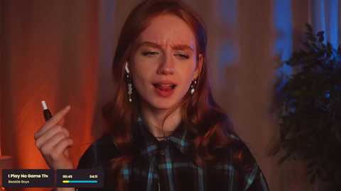 Media: Video of a young woman with fair skin, red hair, and blue plaid shirt, singing passionately, holding a microphone, background blurred with warm lighting and a potted plant.