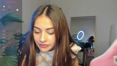 Media: A video of a young woman with long, straight brown hair, wearing a floral shirt, smiling while looking down. She's in a room with a mirror, plants, and a ring light.