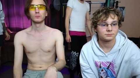 Media: Video of two young men, one shirtless with fair skin and glasses, the other in a hoodie with a graphic design. Background includes a woman in workout clothes and gym equipment.