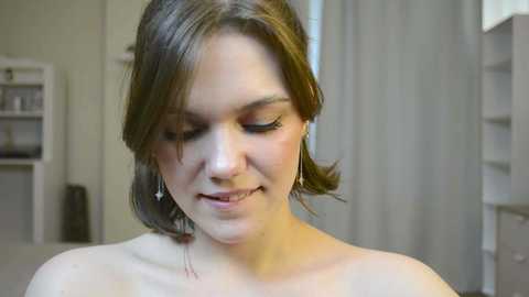 Media: Video of a young woman with short brown hair, wearing a strapless top, standing indoors. She has a fair complexion, and her eyes are closed, smiling gently. The background features a beige wall and white furniture.