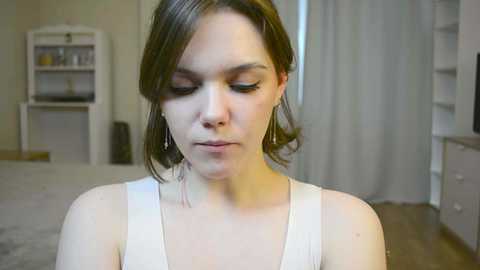 Media: Video of a fair-skinned woman with short brown hair, wearing a white tank top, standing indoors. Background includes beige walls, a white bookshelf, and a white cabinet.