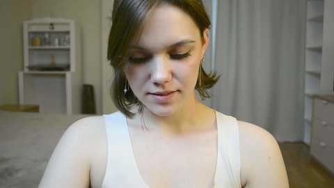 Media: Video of a young woman with fair skin and short brown hair, wearing a white tank top, standing in a softly lit, modern room with neutral tones.